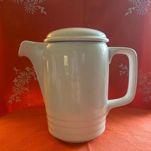 Könnte beinhalten: Eine weiße Keramik-Kaffeekanne mit Deckel, Henkel und Ausguss. Der Topf hat drei rosa Streifen um den Boden. Der Hintergrund ist ein roter Stoff mit weißen Blumenmustern. Die Kanne dient wahrscheinlich zum Servieren von Kaffee oder Tee.
