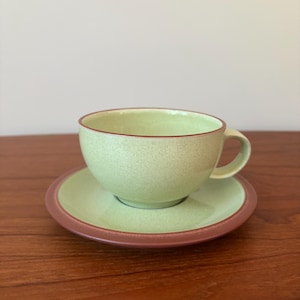 May include: A light green ceramic teacup and matching saucer set. The cup features a speckled finish with a smooth, dark brown rim. The saucer has a matching light green center and a wide, dark brown border, creating a cohesive look.