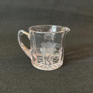 Peut inclure: Crémier en verre transparent avec une anse et un bec verseur. Le crémier présente un motif floral gravé sur le devant et un motif géométrique sur le fond. Le crémier est en verre transparent, sur fond sombre.