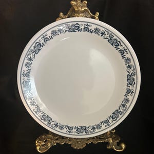 Puede incluir: Plato blanco con un borde floral azul. El plato es redondo y tiene un diseño simple y elegante. El plato se exhibe en un soporte decorativo de color dorado.