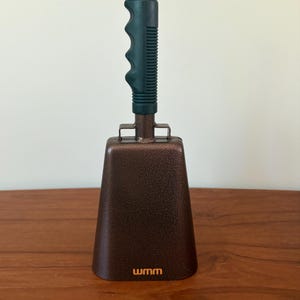 May include: A metallic brown cowbell with a textured finish and a black plastic handle featuring a ridged, ergonomic grip. The lower front of the cowbell displays the letters wmm in a small, orange, sans-serif font centered near the base.