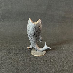 Puede incluir: Pequeña figura de pez de metal negro texturizado con la boca muy abierta. El pez tiene pequeñas aletas y una cola, y está montado sobre una pequeña base redonda de color dorado. La figura está de pie.