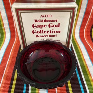May include: A vintage ruby red glass dessert bowl from the Avon Cape Cod Collection, with a scalloped edge. The bowl is presented in front of its original box, which features the text "AVON Bol à dessert Cape Cod Collection Dessert Bowl".