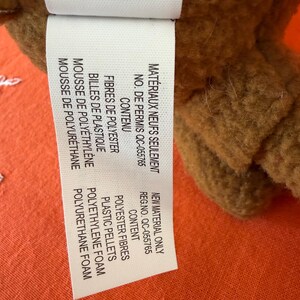 May include: A close-up of a brown plush toy with a white tag. The tag lists the materials: polyester fibres, plastic pellets, polyethylene foam, and polyurethane foam. The tag also includes the text "New Material Only".