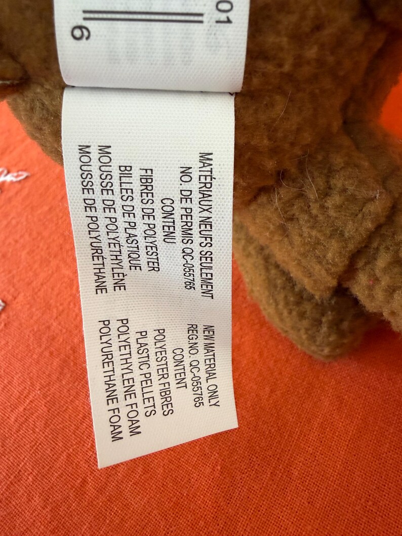 May include: A close-up of a brown plush toy with a white tag. The tag lists the materials: polyester fibres, plastic pellets, polyethylene foam, and polyurethane foam. The tag also includes the text "New Material Only".