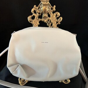 Puede incluir: Un bolso bandolera gris claro con cierre de cremallera y correa larga. El bolso tiene el texto "PIXIE MOOD" impreso en la parte delantera. El bolso se exhibe sobre un soporte decorativo dorado sobre un fondo negro.