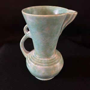 Puede incluir: Una pequeña jarra de cerámica verde claro con base redondeada y parte superior acampanada. La jarra tiene un asa y un pico. La superficie tiene un aspecto moteado. La jarra es probablemente vintage o antigua.