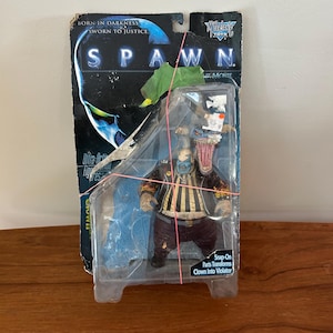 Puede incluir: Figura de acción de Spawn en su embalaje original. La figura es de un payaso con una cara grotesca, que lleva una camisa a rayas y pantalones marrones. El embalaje dice "Spawn The Movie" y "Born in Darkness, Sworn to Justice."