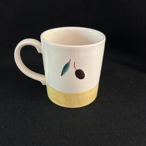 May include: Cream-colored ceramic mug with a yellow base and a curved handle. The mug is decorated with a painted olive branch design, featuring a green leaf and a brown olive. The mug is suitable for hot or cold beverages.