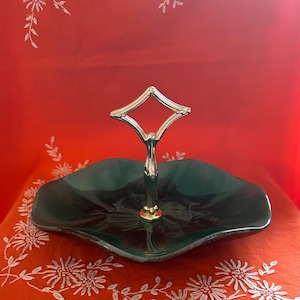 May include: A two-tiered serving tray with a dark green, scalloped dish and a gold-colored handle. The handle has a diamond-shaped top. The tray is on a red surface.