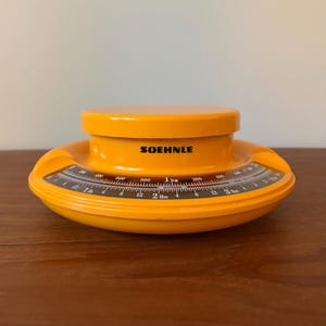 May include: A bright orange Soehnle kitchen scale with a circular platform. The curved display features a black background with white markings, showing measurements in both kilograms up to 1 kg and pounds up to 3 lbs, with clear black text.