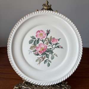 Pode incluir: Um prato de cerâmica branca com um desenho floral de rosas cor-de-rosa e folhas verdes. O prato tem uma borda dourada e uma borda texturizada. É exibido em um suporte decorativo de latão.
