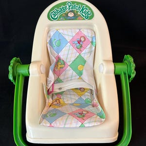 Puede incluir: Un asiento de bebé Cabbage Patch Kids vintage en color crema con detalles verdes. El asiento presenta una tela estampada colorida con personajes de Cabbage Patch Kids. El logotipo de Cabbage Patch Kids está impreso en la parte superior.