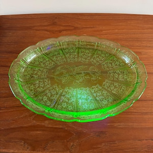 May include: A vintage, oval-shaped, green glass serving tray. The tray has a scalloped edge and is decorated with a floral pattern. The glass has a slight glow, and the tray is resting on a wooden surface.