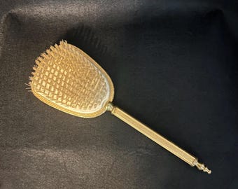 May include: A vintage gold-coloured hairbrush with a rectangular head and a long, slender handle. The brush head features a grid pattern with bristles. The handle has a textured design and a decorative end. The brush is set against a dark background.