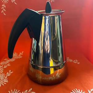 Könnte beinhalten: Ein glänzender Espressokocher aus Edelstahl mit schwarzem Griff und Deckel. Der Kaffeezubereiter spiegelt seine Umgebung wider und steht auf einer orangefarbenen Oberfläche mit weißer Blumenstickerei. Der Hintergrund ist rot.
