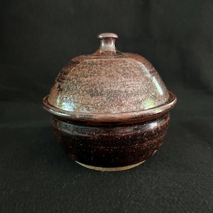 May include: A handmade ceramic bowl with a lid. The bowl and lid are dark brown with a speckled texture. The lid has a small knob. The round bowl sits on a flat base. The design is rustic and earthy, with a natural aesthetic. The bowl is approximately 10 cm in diameter.