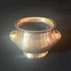 May include: A small, antique silver-coloured bowl with a rounded body and a flared rim. The bowl has two decorative lion head handles and sits on a small, circular base. The interior is dark.