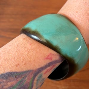 May include: A wide, smooth bangle bracelet featuring a polished finish. The surface displays a gradient colour transition, shifting from a vibrant, bright turquoise green on the outer curve to a deep, solid black along the inner edge and rim.