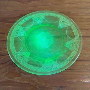 May include: A round, translucent neon green glass plate featuring a central circular embossed pattern and a wide decorative border with repeating geometric and triangular motifs etched into the surface of the glass material.