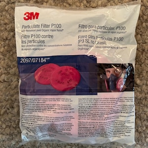 May include: A clear plastic packet containing a 3M Particulate Filter P100. The packet features text in English, French, and Spanish, and contains two red circular filters. The product is designed for respiratory protection.