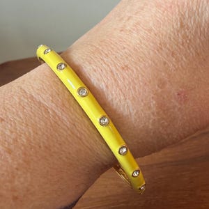Op de afbeelding: Een felgele emaille armband met een reeks gelijkmatig verdeelde, ronde, heldere stenen die in goudkleurige metalen zettingen aan de buitenkant van de band zijn geplaatst. De armband heeft een gladde, glanzende afwerking en een scharniersluiting.