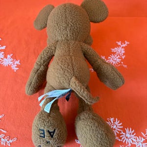 May include: A brown mouse-shaped plush toy, seen from the rear. It features large ears, a tail, and the letters "AE" on one foot. The toy is made from a soft, textured fabric.