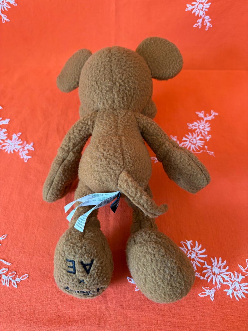 May include: A brown mouse-shaped plush toy, seen from the rear. It features large ears, a tail, and the letters "AE" on one foot. The toy is made from a soft, textured fabric.