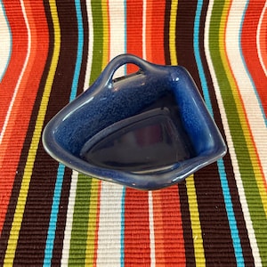 Könnte beinhalten: Eine kleine, dreieckige blaue Keramikschale mit Griff. Die Schale hat eine glänzende Oberfläche und eine gesprenkelte Textur. Der Hintergrund ist ein farbenfrohes gestreiftes Gewebe in Rot-, Orange-, Braun-, Grün-, Blau-, Gelb- und Weißtönen.