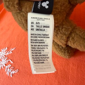 May include: A brown plush toy with a Disney tag. The tag reads "AMERICAN EAGLE OUTFITTERS" and includes the text "US O/S". The tag also lists the materials used in the toy's construction.