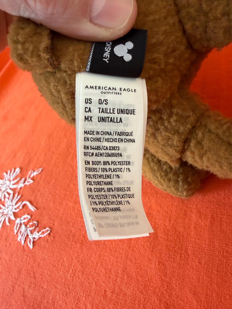 May include: A brown plush toy with a Disney tag. The tag reads "AMERICAN EAGLE OUTFITTERS" and includes the text "US O/S". The tag also lists the materials used in the toy's construction.