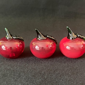 以下が含まれることがあります： 黒い茎と葉が付いた、3つの小さな赤いガラス製のリンゴの置物。リンゴは光沢があり、光を反射し、暗い表面に置かれています。ガラス製のリンゴは装飾品です。