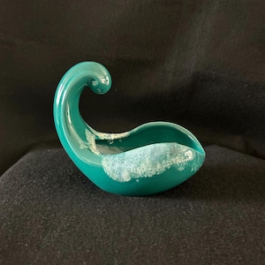 Puede incluir: Un cuenco de cerámica de color turquesa con un glaseado blanco. El cuenco tiene forma de ola y tiene un asa en un lado.