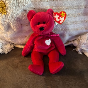 Può includere: Un orsacchiotto di peluche rosso con un cuore bianco sul petto. L'orso ha un naso nero e un nastro rosso. Un'etichetta rossa a forma di cuore con la scritta "ty" è attaccata all'orso.