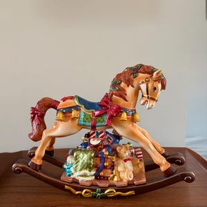 May include: A decorative rocking horse figurine in shades of tan, brown, and red. The horse is adorned with festive details, including a blue saddle, red ribbons, and gold bells. A collection of wrapped gifts and a teddy bear are at the base.