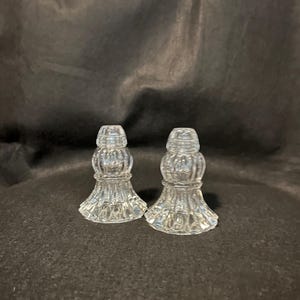 May include: Two clear, vintage-style glass salt and pepper shakers. Each shaker has a bulbous body with vertical ribbing and a flared base. The tops are also ribbed and have a small opening for dispensing. The shakers are displayed against a dark background.
