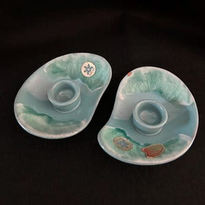May include: A pair of turquoise ceramic candle holders with an irregular, organic form. Each has a central well for a candle and a decorative, wave-like pattern in green and white. Small stickers are attached to the surface of each holder.