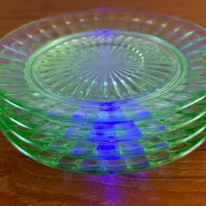 Può includere: Una pila di quattro piatti di vetro verde. Ogni piatto ha un bordo a coste e un design centrale strutturato. I piatti sono impilati uno sopra l'altro, con una luce blu che li attraversa.