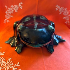 Peut inclure: Figurine de grenouille en céramique vert foncé brillant, avec un corps marron foncé et une rayure blanche sur le dos. La grenouille a des pieds palmés, sur fond orange vif.