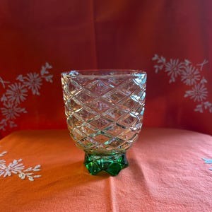 Może przedstawiać: Przezroczysty szklany kubek z rombowym wzorem, spoczywający na zielonej podstawie. Szkło ma teksturowaną powierzchnię i zostało zaprojektowane tak, aby wychwytywać światło. Kubek to szklanka do picia w stylu vintage.
