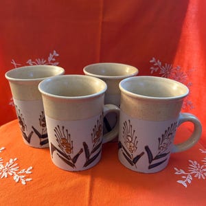 Peut inclure: Quatre mugs en céramique avec un bord beige et une partie inférieure blanche. Chaque mug présente un motif floral marron et noir. Les mugs sont sur une surface orange avec une broderie florale blanche et un fond rouge.