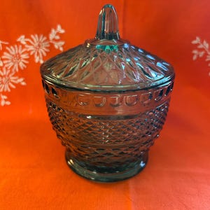 May include: A teal glass candy dish with a lid. The dish features a diamond pattern on the body and lid, and a small handle. It is set on an orange surface.