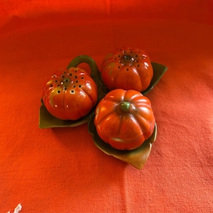 May include: A set of three orange ceramic pumpkin-shaped salt and pepper shakers. Each shaker has a green stem and sits on a green leaf-shaped base. The shakers are arranged on an orange tablecloth.