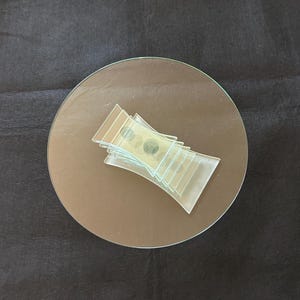 Può includere: Una pila di vetrini trasparenti per microscopio con campioni, appoggiata su una superficie rotonda, riflettente e marrone. I vetrini sono disposti a ventaglio, su uno sfondo di tessuto scuro e strutturato.