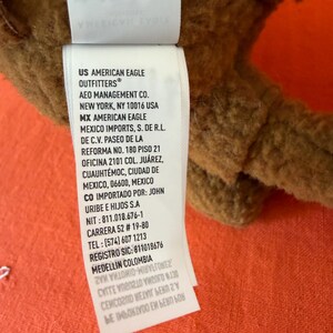 May include: A brown plush toy with a white tag, featuring text in English and Spanish. The tag includes the brand "American Eagle Outfitters" and care instructions. The background is an orange textured fabric.