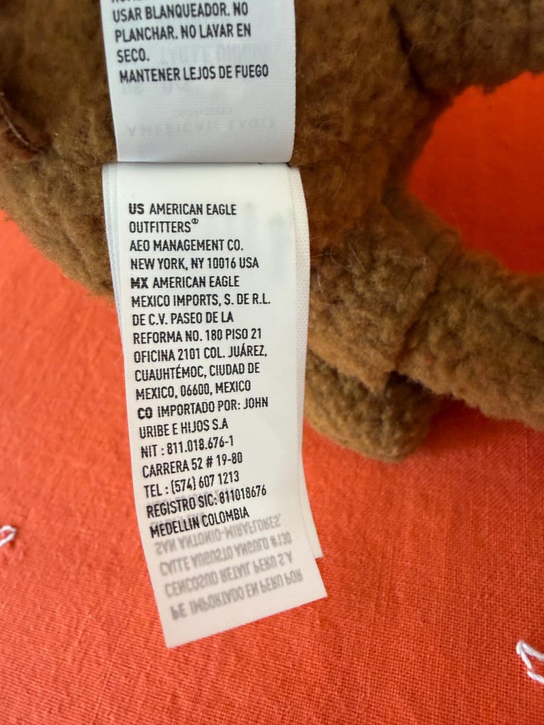 May include: A brown plush toy with a white tag, featuring text in English and Spanish. The tag includes the brand "American Eagle Outfitters" and care instructions. The background is an orange textured fabric.