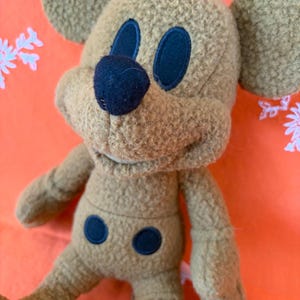 May include: A tan-coloured plush toy resembling a cartoon mouse, with dark blue eyes, nose, and belly details. The toy features large, rounded ears and a soft, textured finish. Set against a bright orange backdrop.