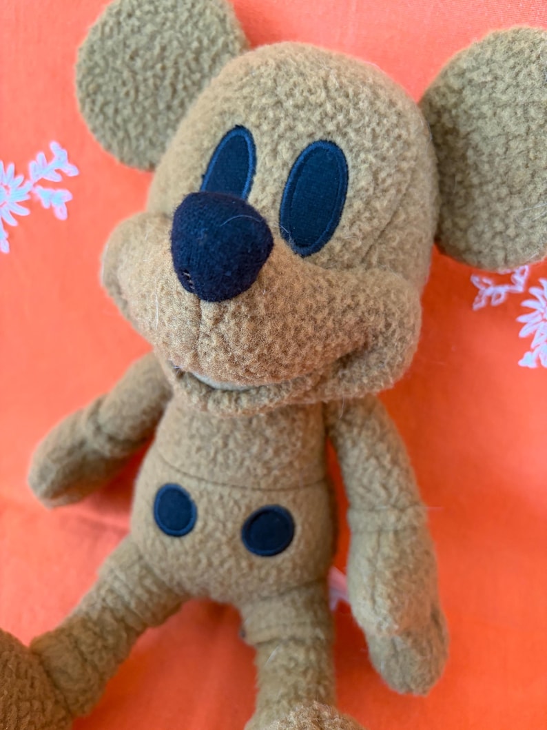 May include: A tan-coloured plush toy resembling a cartoon mouse, with dark blue eyes, nose, and belly details. The toy features large, rounded ears and a soft, textured finish. Set against a bright orange backdrop.