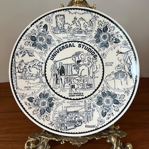 Op de afbeelding: Een decoratieve bord met scènes uit Universal Studios California, met tekst als "Naval Battle on Lagoon" en "Glamor Tram Through European Street". Het bord heeft een witte achtergrond met zwarte illustraties en bloemaccenten, op een goudkleurige standaard.