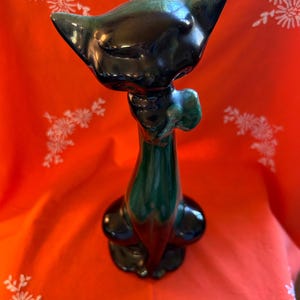 May include: A glossy black and green ceramic cat figurine. The cat has a long neck, a bow tie, and a sleek, stylized design. The figurine is set against a red background.